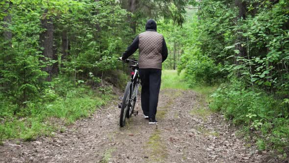 Man Walks and Rolls a Bicycle Next to Him Along a Forest Path alt