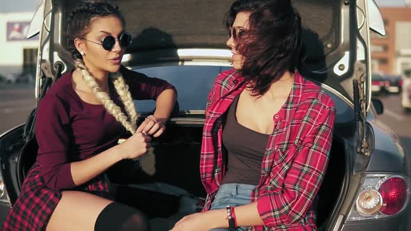 Two Young Attractive Women in Stylish Sunglasses Sitting in Car Trunk in the Parking By the Shopping alt