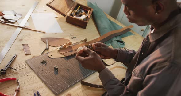 Focused african american craftsman using tools to make a belt in leather workshop alt