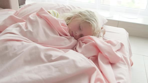 Adorable Blonde Girl Sleeping Sweetly in the Morning on Pink Bed Linens at Home alt