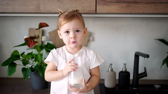 Happy Baby Girl Sitting at the Table in the Kitchen and Drinking Milk alt