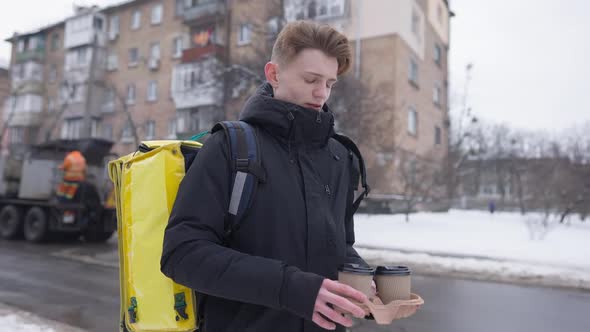 Thoughtful Young Man Standing with Yellow Insulated Thermal Backpack and Disposable Coffee Cups alt
