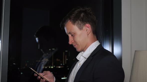 Young Businessman in Office Suit Is Browsing Smartphone Near the Window with City View. alt