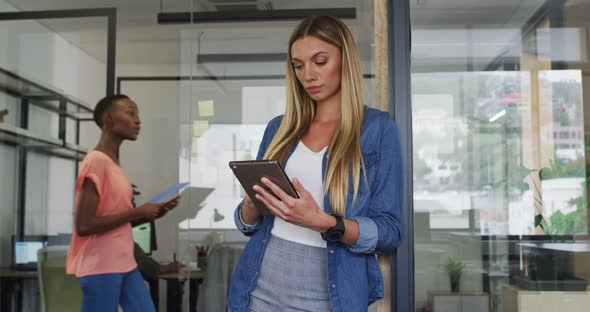 Caucasian businesswoman leaning in doorway using tablet, smiling to camera in busy office alt