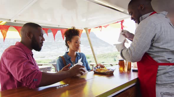 African american man wearing apron serving burgers and fries to a couple at the food truck alt