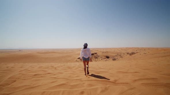 Young Woman in Jeans and White Shirt Walking Down Sand Dune in the Desert alt