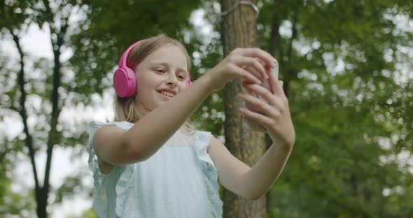 Teenager Listens to Music on Pink Headphones in Park Making Online Video Call alt
