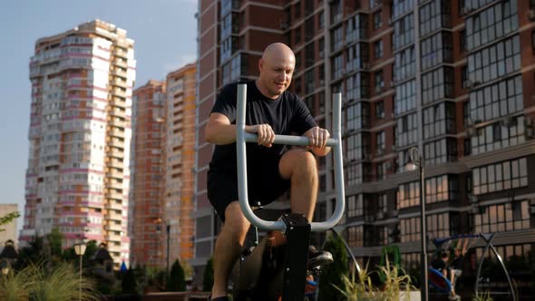A Bald Man Trains on a Stationary Exercise Bike in the Courtyard of a Residential Complex