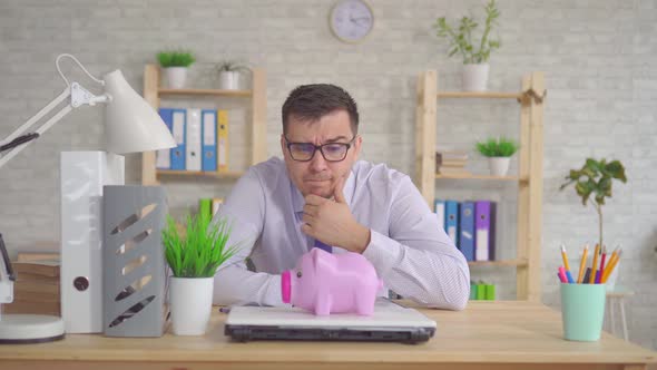 Young Man in a Shirt and Glasses Sitting in the Office Puzzled Looks at the Piggy Bank alt