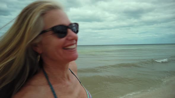 Closeup of laughing, playful mature woman in bikini and sunglasses turns around and runs on the beac alt