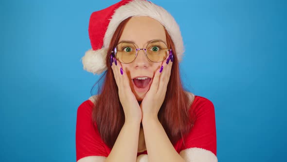 Woman in santa costume opens her mouth in surprise, leaning hands against her cheeks. alt