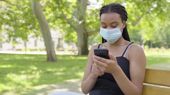 A Young Black Woman in a Face Mask Works on a Smartphone and Sits on a Bench in a Park alt
