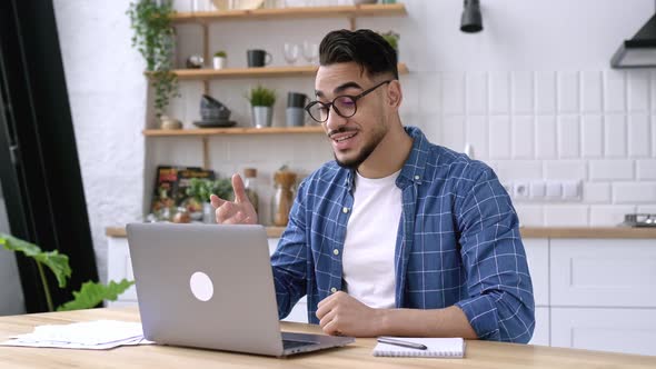 Friendly Happy Arabian or Indian Man with Glasses Sitting at Home in the Kitchen Conducts Video Call alt