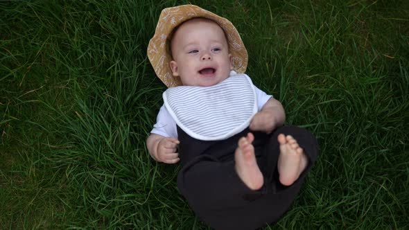 Small Happy Newborn Child in Summer Panama Hat Fall Down Laying on Grass Barefoot in Summer Sunny