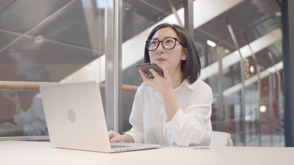 Asian Businesswoman Recording Voice Message on Workplace alt
