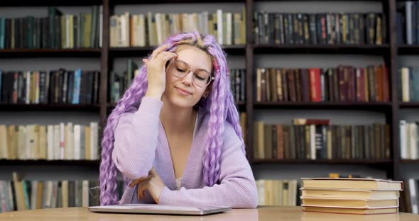 Portrait of a Young Woman with Long Colored Purple Hair Sitting at a Desk with a Laptop and Books in