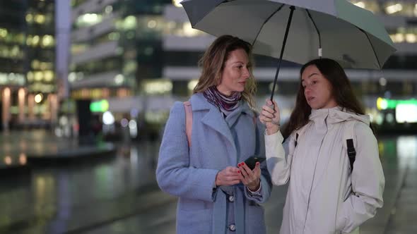Two Women Stand Under an Umbrella and Chat Against the Background of a Blurred Evening Street and alt