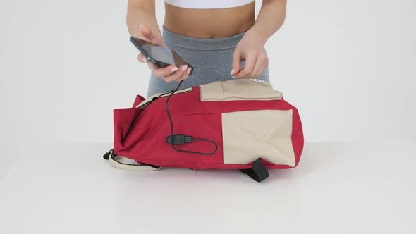 Young woman in gray leggings connects her phone to USB charging port in red backpack