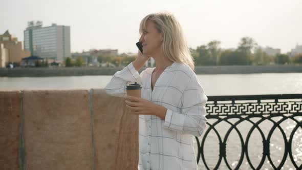 Cheerful Blond Lady Chatting on Mobile Phone Drinking Coffee Walking Along River Embankment in City alt