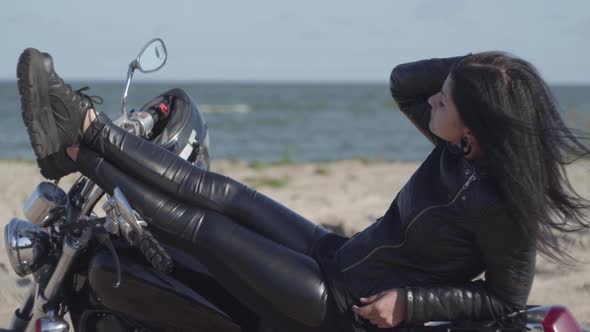 Pretty Young Caucasian Woman in a Black Leather Jacket and Pants Lying on a Motorcycle Near Sea alt