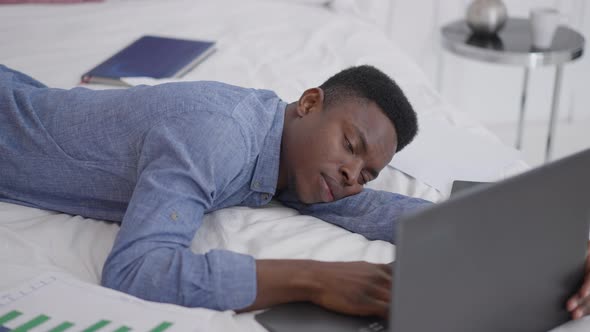 African American Young Businessman Falling Asleep Lying on Bed with Laptop alt