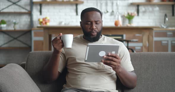 African Man Watching Video on Tablet PC and Drinking Coffee While Sitting on Sofa at Home alt