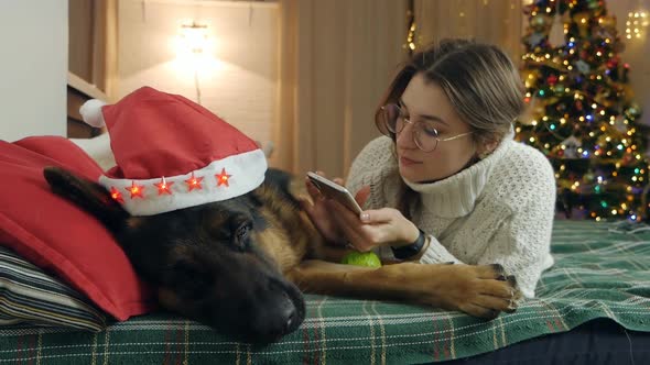 Young Woman Spend Christmas Holidays at Home with Her Pet on the Bed with Phone