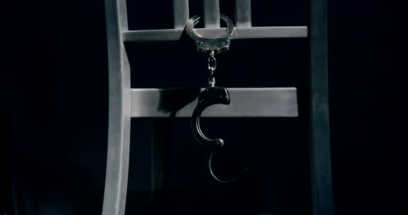 Medium tracking shot of a pair of handcuffs hanging from the back of a chair. The cuff dangling down alt