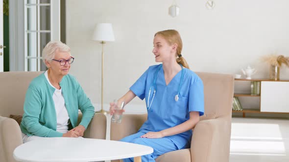 Positive medical worker in uniform carries glass of clean water to mature lady alt