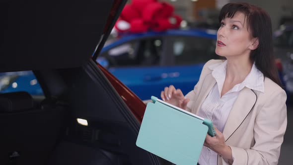 Professional Serious Female Car Dealer Examining New Luxurious Automobile in Dealership Using Tablet alt