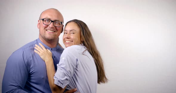 Cheerful Spouses Are Smiling Broadly To Camera and Embracing on White Wall Background alt