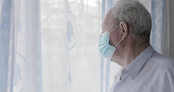 Sad Senior Man in Medical Mask Looking Out the Window and Around with Fear alt