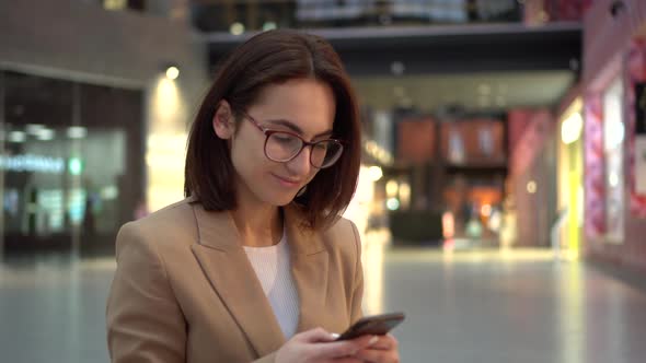 A Young Woman Sits with a Phone in Her Hands in a Shopping Center with a Large Paln. A Girl in a alt