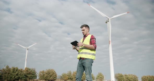 Young Engineer with Tablet in Hand Looks Behind and Works on Electric Turbines alt