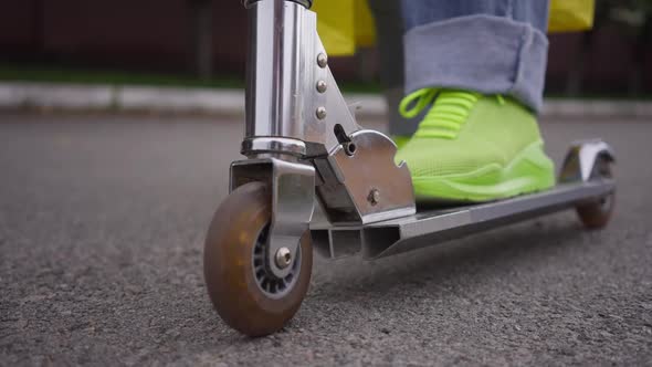 Closeup Female Foot in Yellow Green Sneakers and Denim Jeans Standing on Retro Kick Scooter in Slow alt