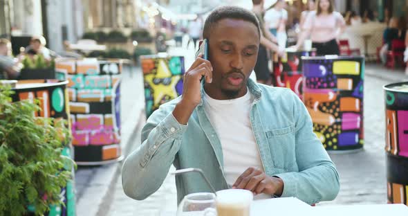 Man in Casual Clothes with Stylish Beard which Sitting in Cozy Street Cafe and Talking on Phone alt