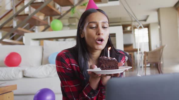 Mixed race woman using laptop having birthday video chat holding a cake blowing candle alt