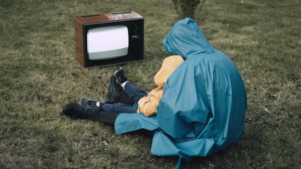 Woman with Small Child in Raincoats are Sitting on Grass and Watching Retro TV