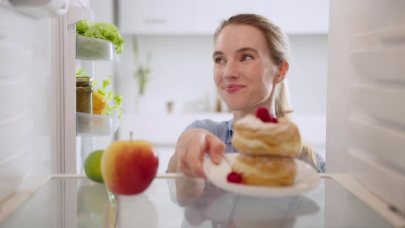 Woman taking cake choosing between an apple from refrigerator. View from inside the working fridge.