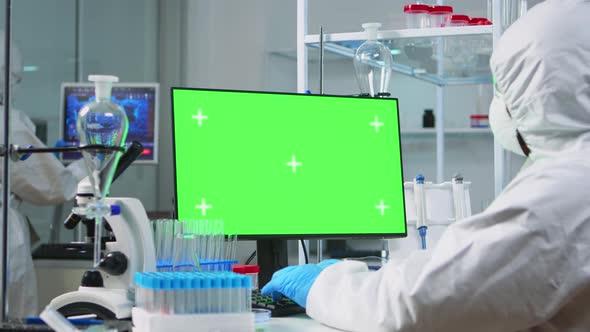Doctor Wearing Protection Suit Typing on Computer with Green Mockup alt