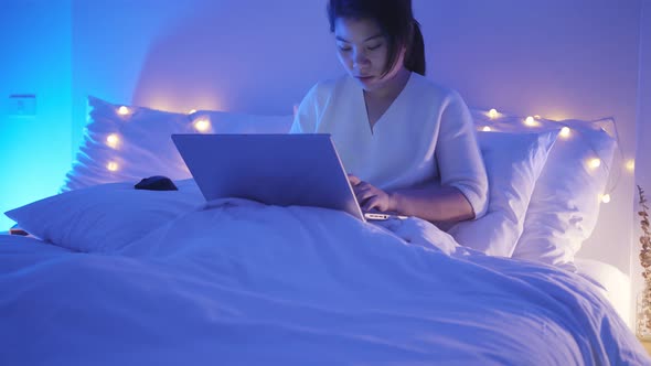 young asian female wearing casual dress sit on bed with meeting online via laptop