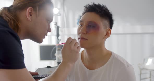 Male Visagiste Applying Bruise Makeup on Face of Young Man Before Shooting Scene alt
