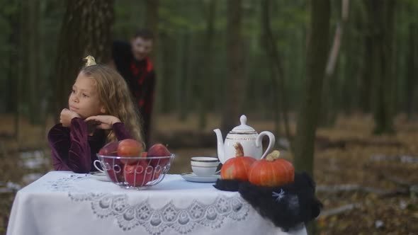 Portrait of Pretty Caucasian Girl Sitting at Table in Forest on Halloween with Blurred Vampire alt