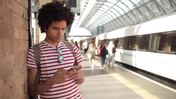 Man at train station checking timetables on mobile phone alt