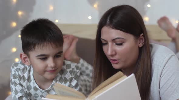 A Beautiful Mother with Long Hair Reads a Book to a Small Boy Lying on a Bed and Kisses Him Having a alt