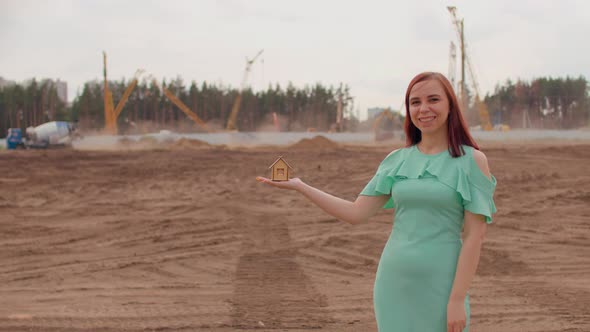 Young Woman with Small Wooden House in Hand Stands on Construction Site alt