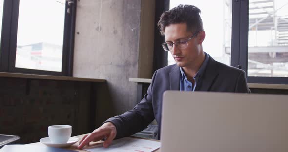 Caucasian businessman using laptop looking at paperwork, sitting at table with coffee in cafe alt