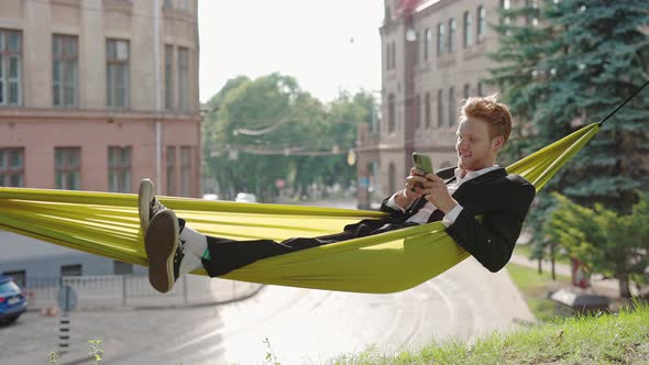 Smiling Businessman Wearing Formal Suit and Casual Sneakers Typing Messages on Smartphone While alt