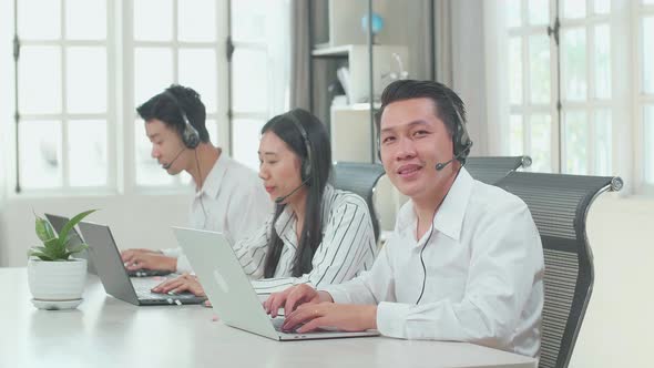 A Man Of Three Asian Call Centre Agents Smiling To Camera While His Colleagues Speaking To Customers alt