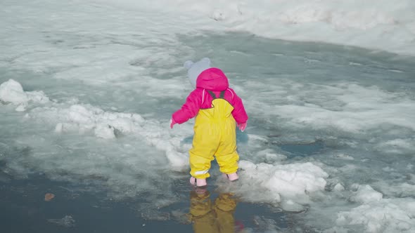 Toddler runs through puddles melting snow in a yellow rubber jumpsuit. Top view alt